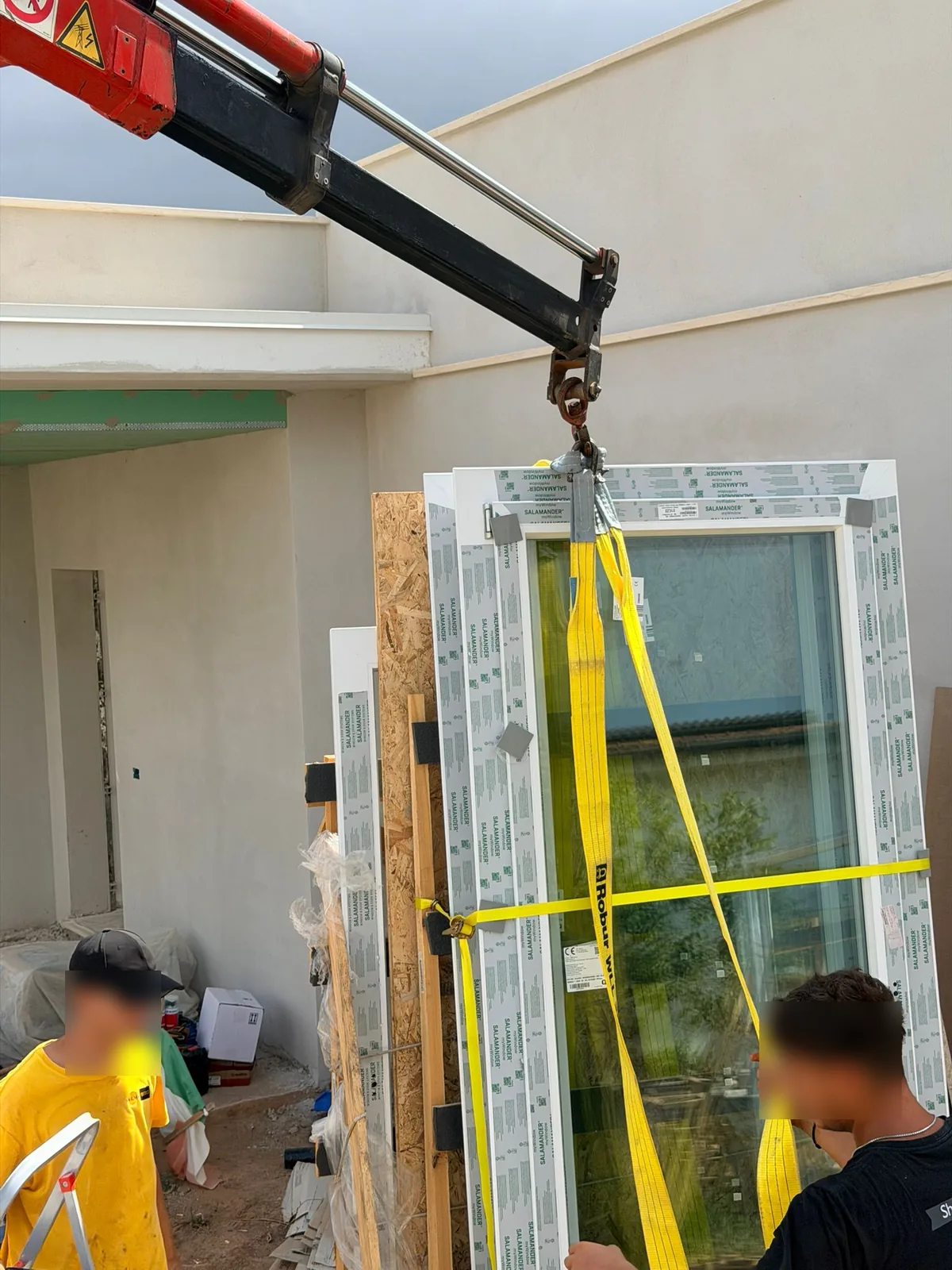 Autogru che solleva una portafinestra imballata con fasce gialle in cantiere, due operai la accompagnano