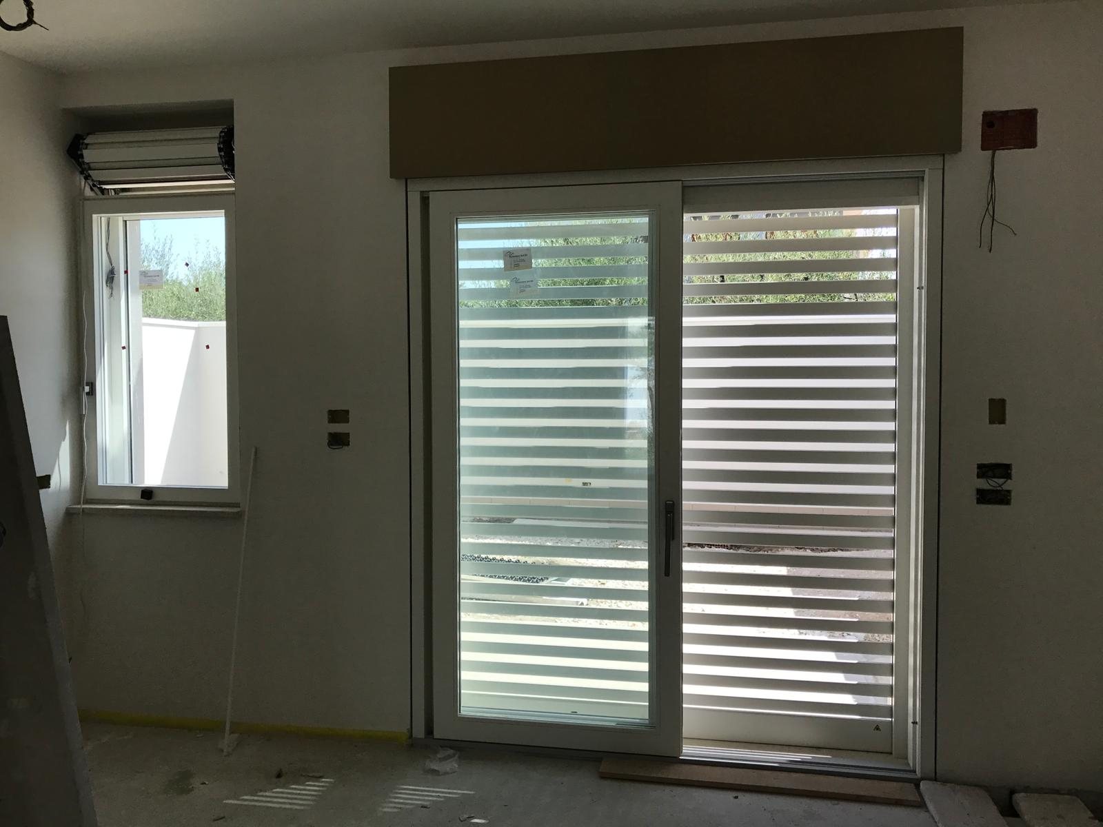 Tapparella blindata Brisolè bianca a doghe distanziate dietro porta-finestra scorrevole in cantiere, vista dall'interno