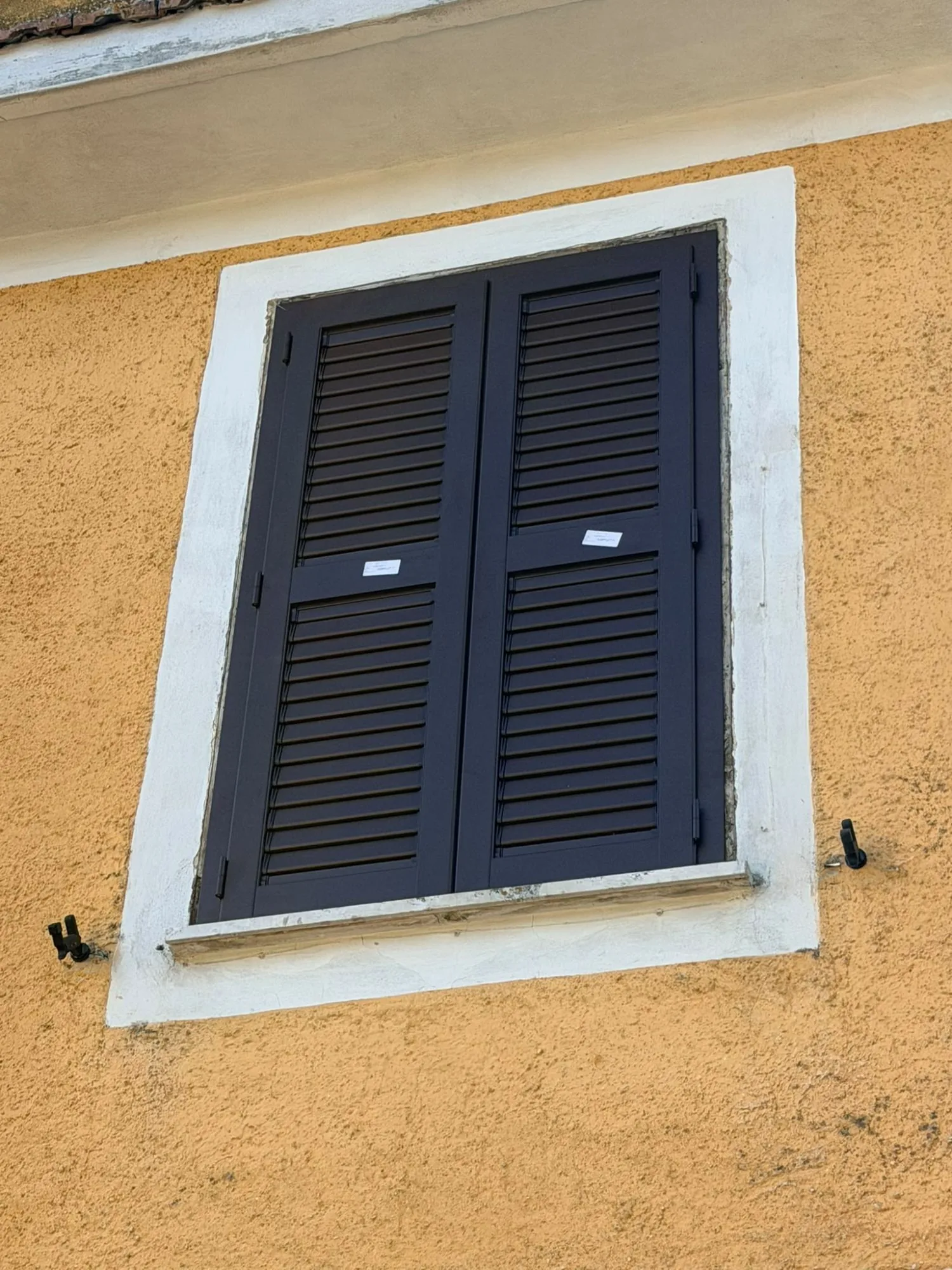 Persiana in alluminio marrone scuro a due ante chiusa, con etichette di produzione, in cornice bianca su facciata gialla