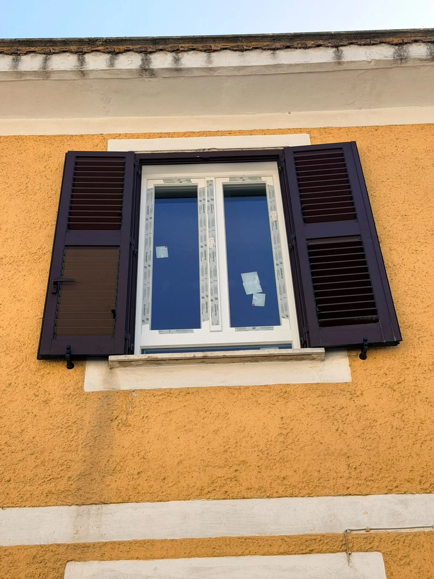Finestra in PVC bianco a due ante fra due persiane in alluminio marrone, su una facciata gialla