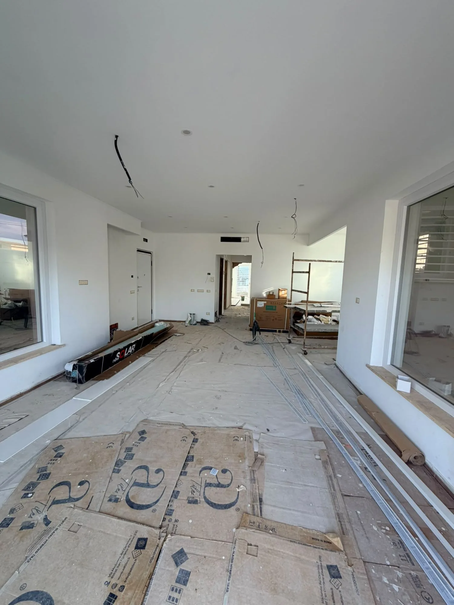 Soggiorno di una casa in cantiere con due grandi finestre fisse in PVC bianco, pavimento coperto da cartoni