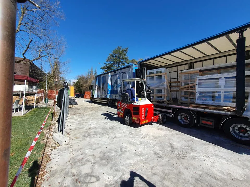 Autoarticolato con serramenti imballati su cavalletti di legno e carrello elevatore rosso che scarica in cantiere