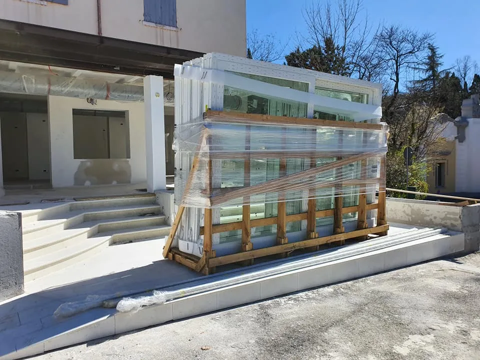 Cassa in legno con serramenti in PVC bianco imballati davanti a un edificio in ristrutturazione con scalinata in pietra