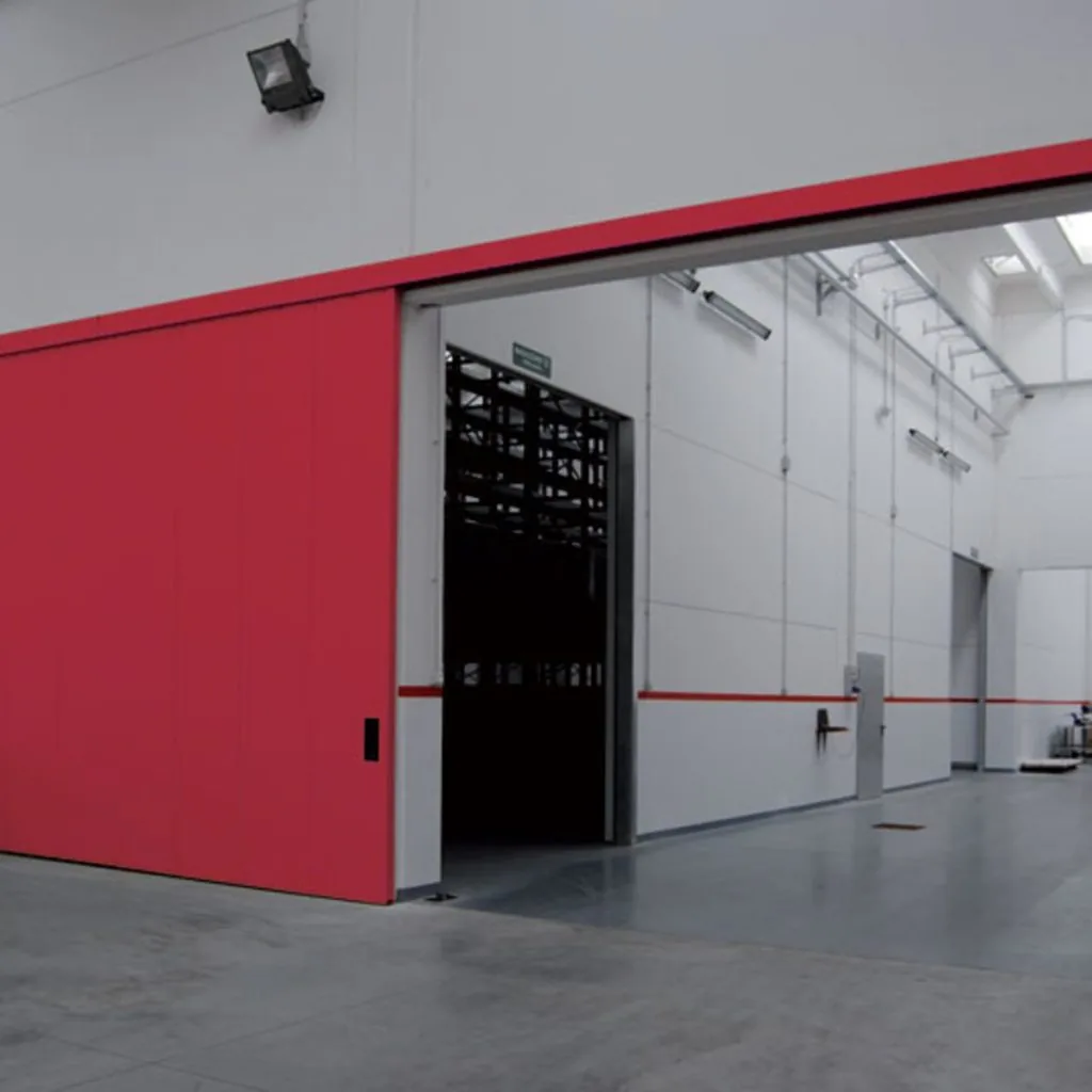 Portone scorrevole rosso a pannelli, aperto a metà, sul passaggio verso un magazzino con scaffalature.