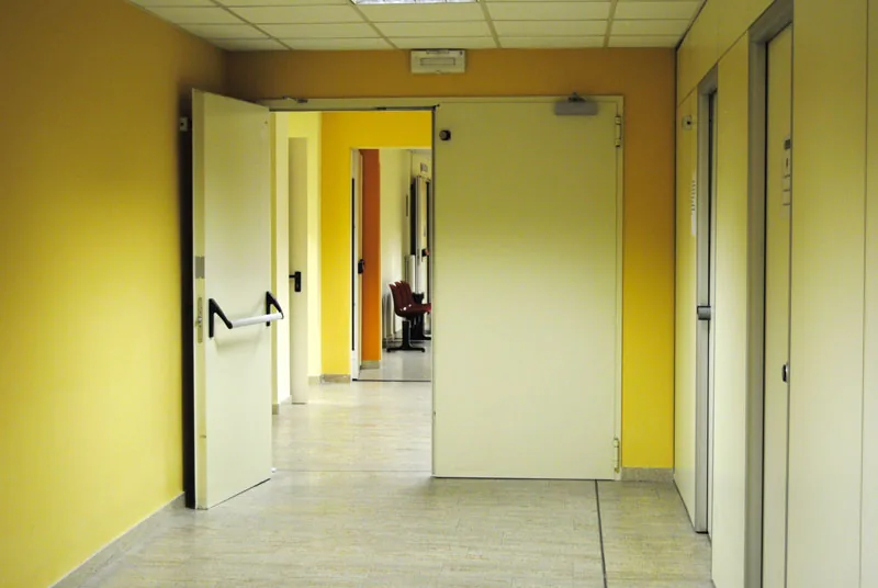 Corridoio con una porta tagliafuoco a due ante: un'anta aperta con maniglione antipanico, l'altra chiusa.