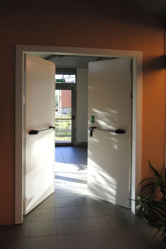 Porta bianca a due ante aperte con telaio abbracciante e maniglioni, parete arancione, luce dal giardino.