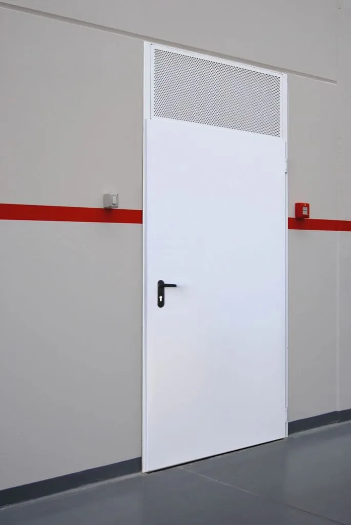 Porta metallica bianca a un'anta con maniglia nera e, sopra, un sopraluce in lamiera forata, in un capannone.