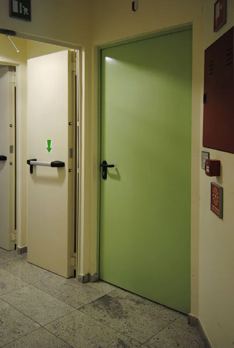 Porta metallica verde a un'anta con maniglia nera; a sinistra una porta color avorio aperta con maniglione antipanico.