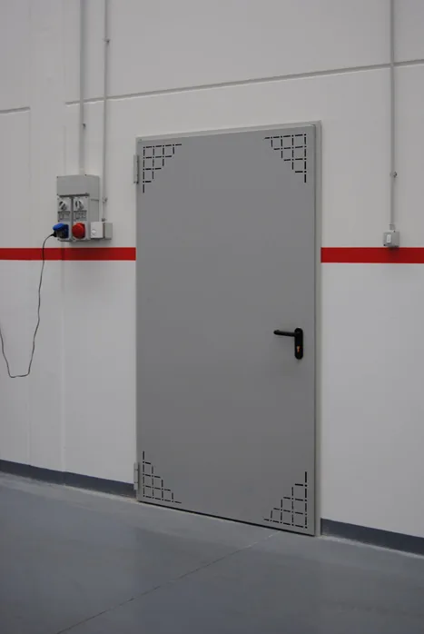 Porta metallica grigia a un'anta con forature di aerazione triangolari ai quattro angoli, in un capannone.