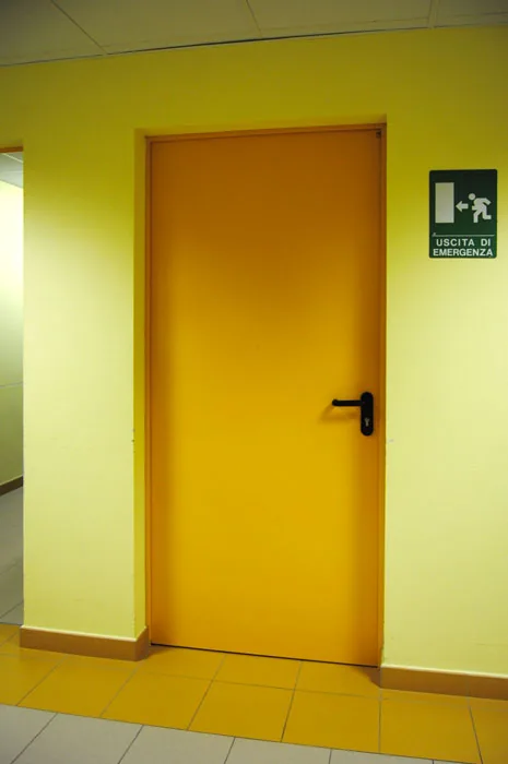 Porta metallica arancione a un'anta con maniglia nera, in un corridoio giallo accanto a un segnale di uscita.