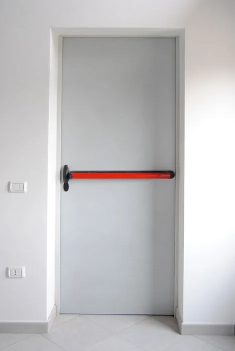 Porta metallica grigio chiaro a un'anta, chiusa, con maniglione antipanico rosso, su una parete bianca.
