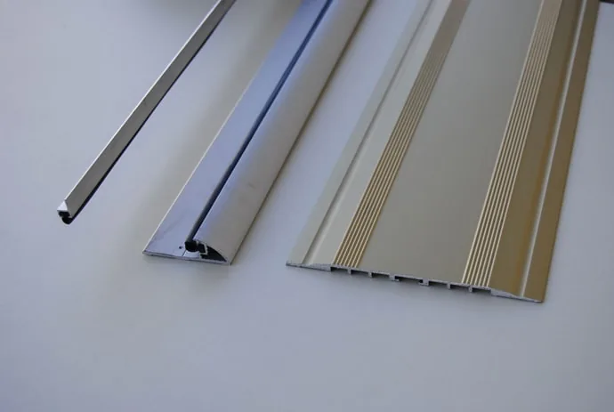 Tre profili in alluminio appoggiati su un piano: una guarnizione sottile, un profilo con guarnizione e una soglia rigata.