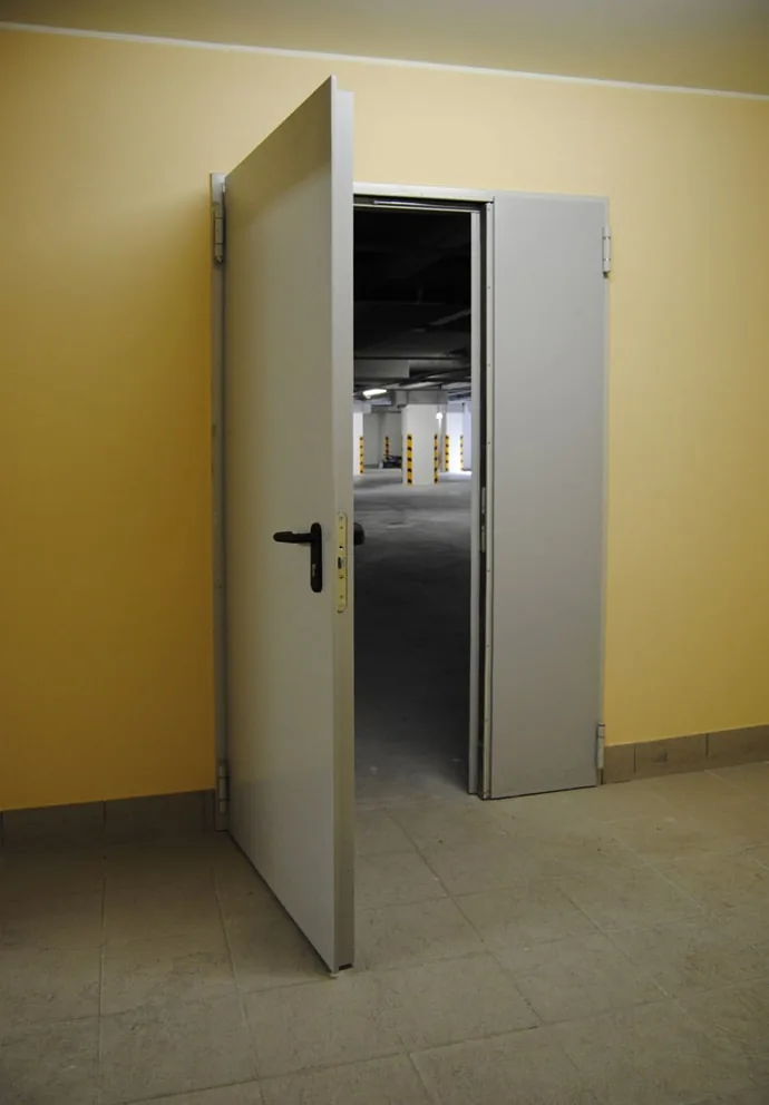 Porta tagliafuoco metallica grigia a due ante, con l'anta principale aperta su un'autorimessa interrata con pilastri.