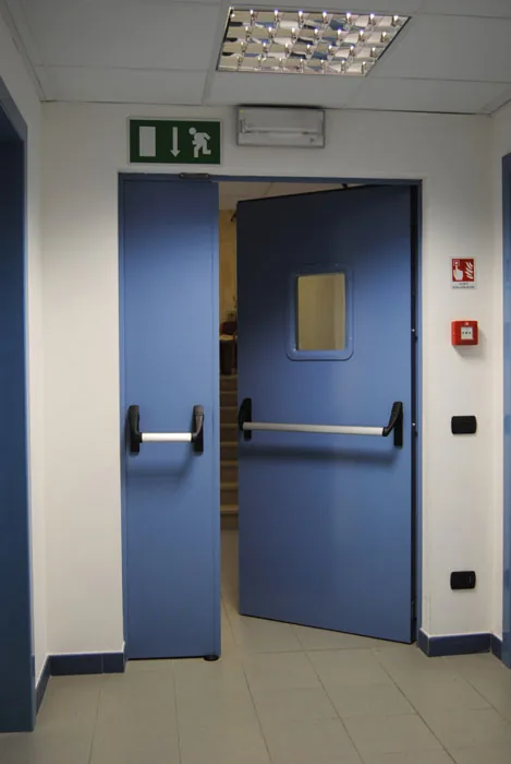 Porta tagliafuoco a due ante con oblò rettangolare e maniglioni antipanico, socchiusa verso un vano scala.
