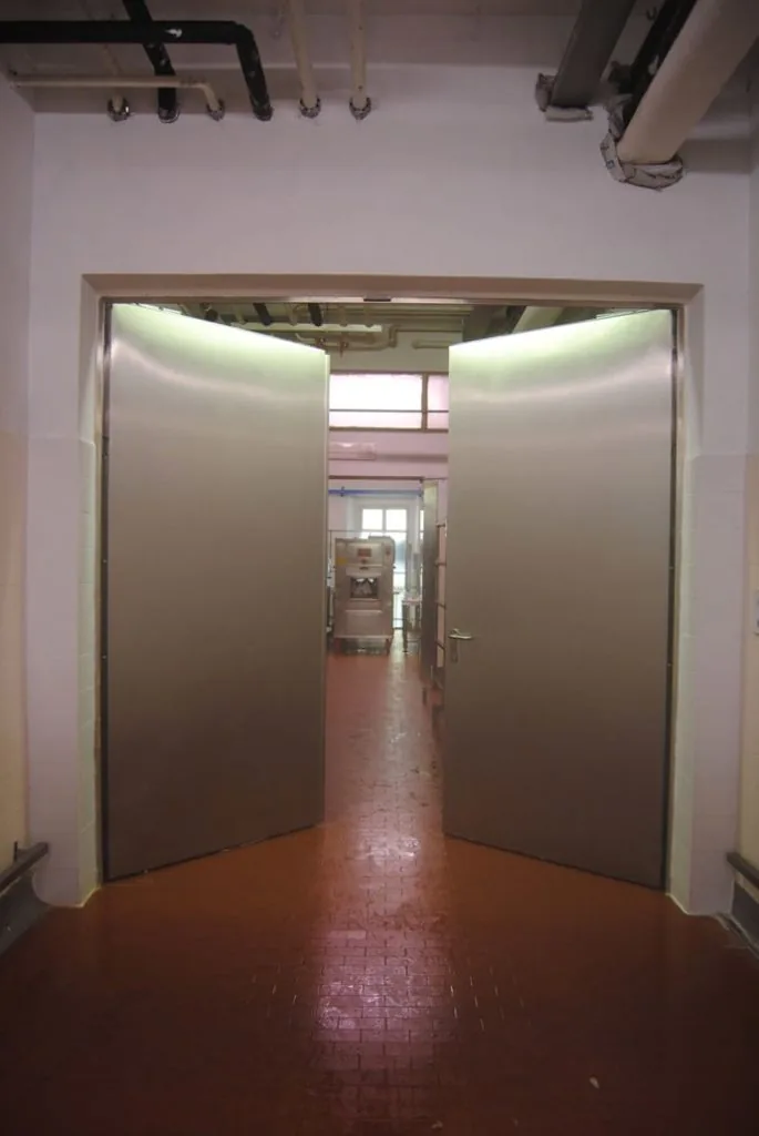 Porta tagliafuoco in acciaio inox a due ante, entrambe aperte, in un ambiente di lavorazione con pavimento in cotto.