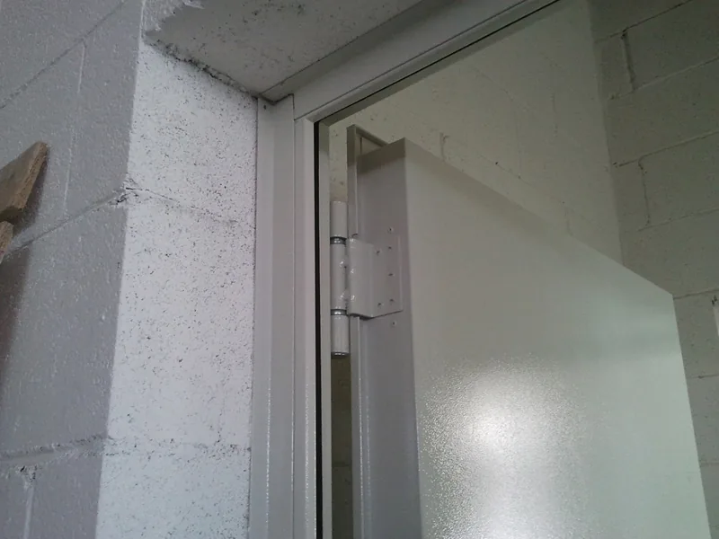 Angolo superiore di una porta metallica aperta: telaio fissato a un muro in blocchi di cemento e cerniera dell'anta.