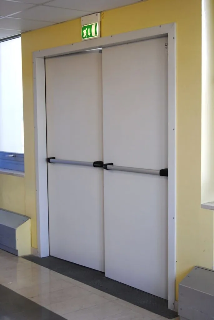 Porta tagliafuoco a due ante chiusa, con telaio abbracciante e due maniglioni antipanico, sotto un segnale di uscita.