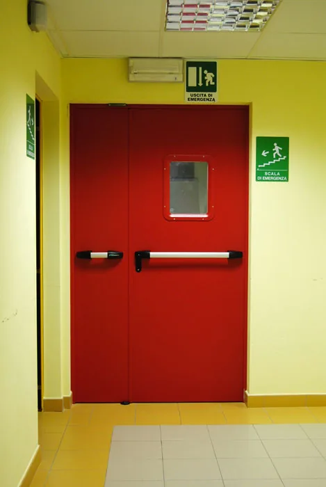 Porta tagliafuoco rossa a due ante chiusa, con oblò rettangolare sull'anta principale e maniglioni antipanico.