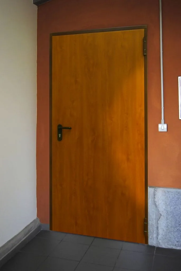 Porta a un'anta con rivestimento effetto legno, telaio scuro e maniglia nera, su una parete color mattone.