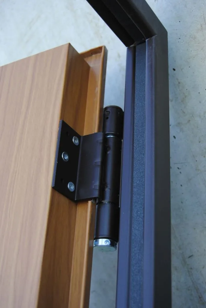 Cerniera nera che unisce un'anta rivestita effetto legno al telaio verniciato grigio scuro, vista con la porta aperta.