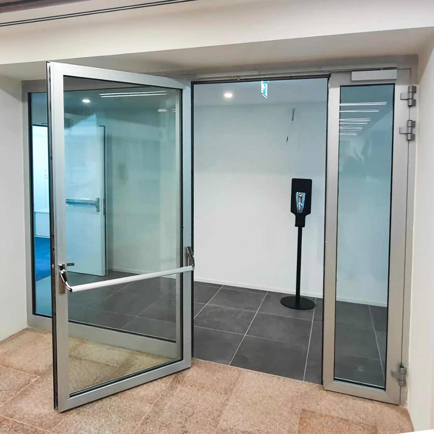 Porta vetrata tagliafuoco a un'anta aperta, con maniglione antipanico e fisso laterale, profili in alluminio argento.