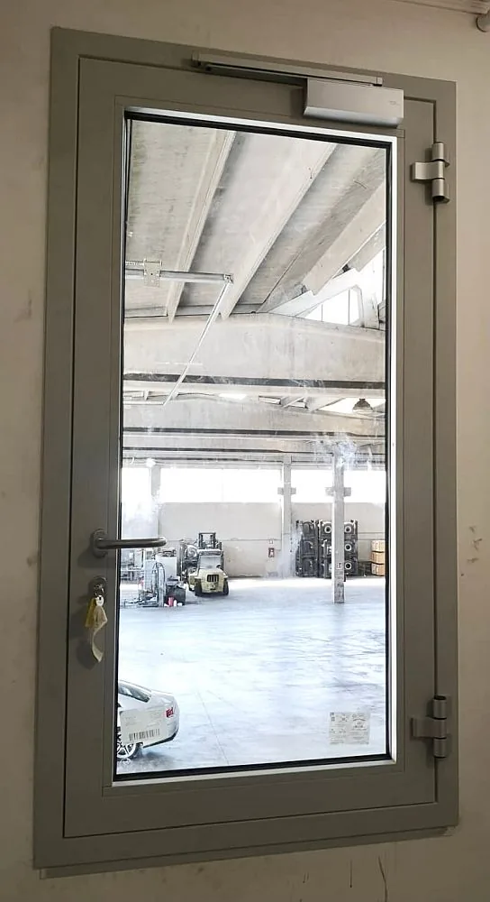 Porta vetrata a un'anta con profili in alluminio e chiudiporta; oltre il vetro, un capannone con carrelli elevatori.