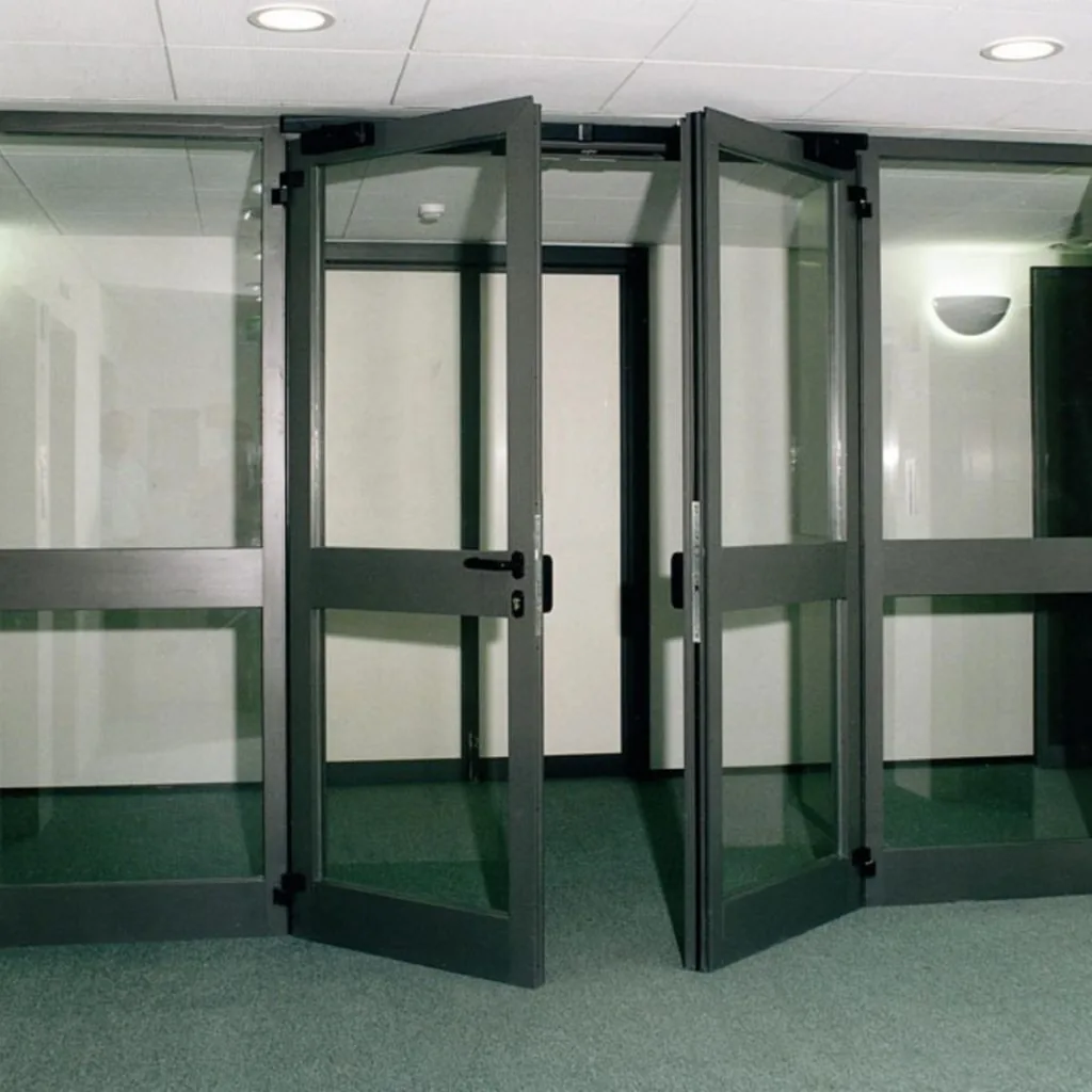 Vetrata tagliafuoco con profili scuri: porta a due ante aperte tra due specchiature fisse, in un atrio con moquette.