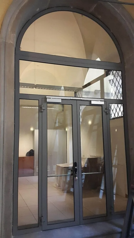 Vetrata ad arco con profili antracite: porta a due ante, fianchi vetrati e sopraluce dentro un portale in pietra.