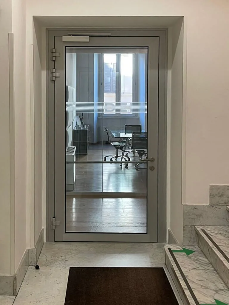 Porta vetrata a un'anta con profili in alluminio, chiudiporta aereo, tre cerniere, maniglione e fascia satinata sul vetro.