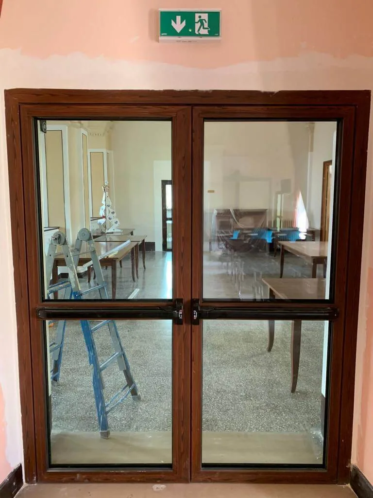 Porta vetrata a due ante con profili effetto legno scuro e maniglioni antipanico neri, in un salone in ristrutturazione.