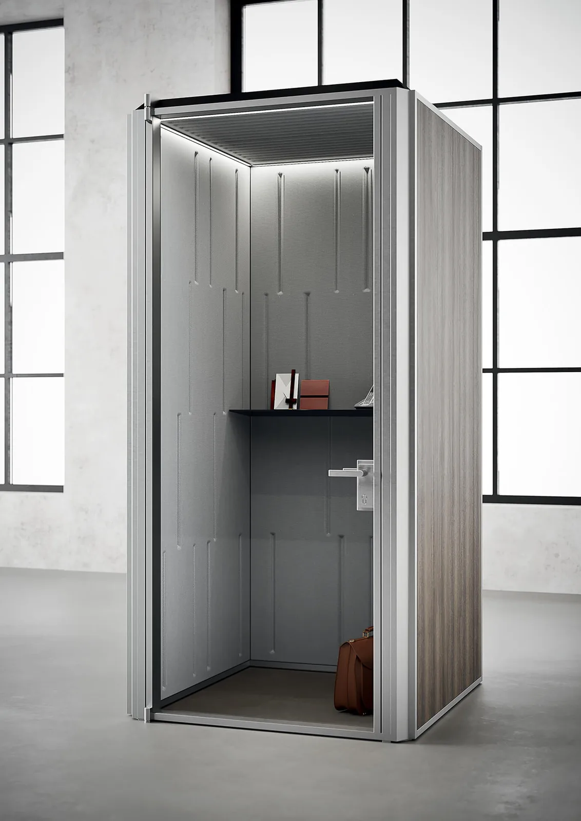 Cabina acustica per una persona con porta in vetro e soffitto con luce, in un ambiente ampio con pavimento in cemento e grandi finestre